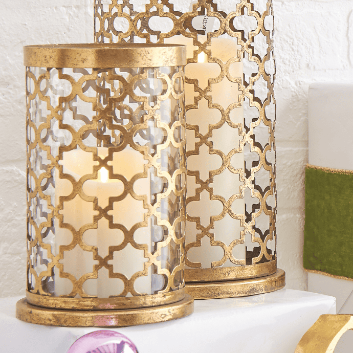 Two decorative candle holders with intricate gold lattice patterns surrounding white pillar candles, placed on a white surface. A velvet green accent and a pink ornament are visible nearby.