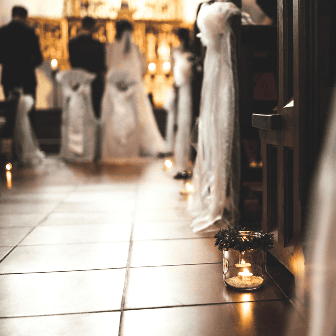 A softly lit church aisle is decorated with small glass jars containing candles and greenery. In the blurred background, a couple stands at the altar with guests seated on either side, draped in white fabric.