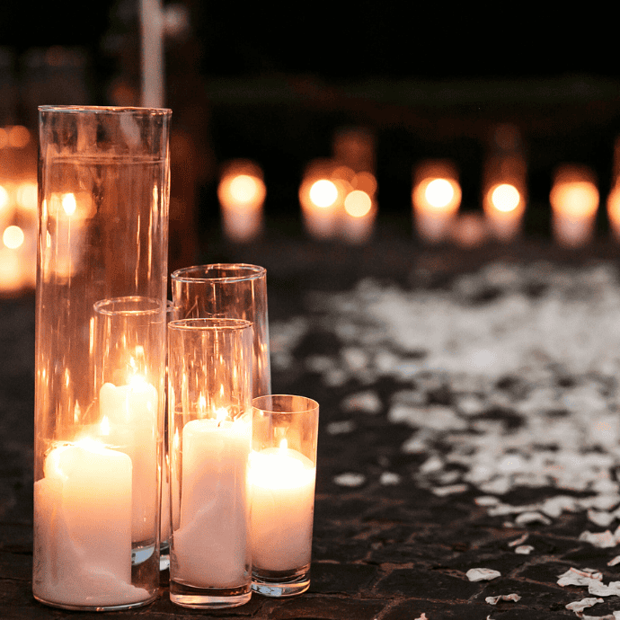 Tall glass vases with lit candles are placed on a dark surface surrounded by scattered white rose petals. In the background, more candles softly illuminate the scene, creating a warm and intimate atmosphere.