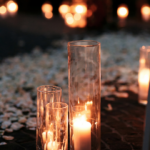 Tall glass vases with lit candles inside are placed on a stone path, surrounded by scattered flower petals. The scene is softly illuminated by the warm glow of candlelight, creating a cozy and intimate ambiance.