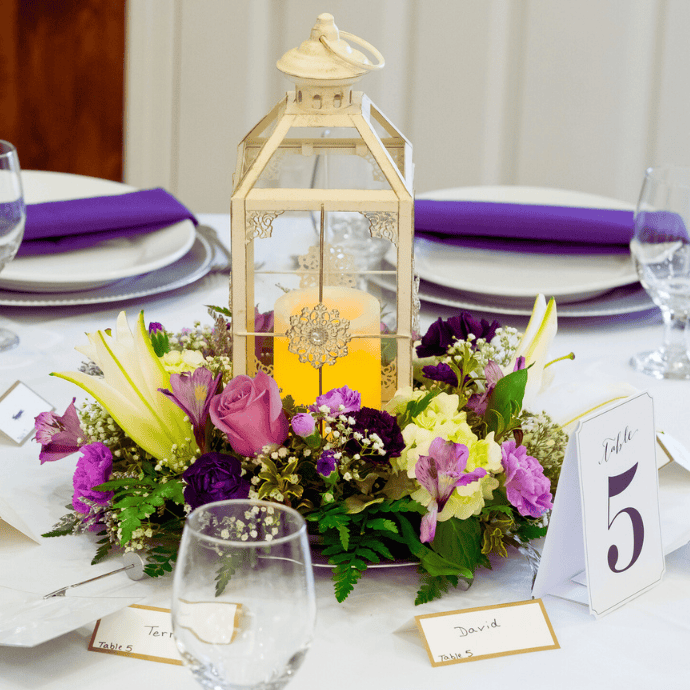 A decorative lantern with a glowing candle is surrounded by purple and yellow flowers on a white table. Plates have purple napkins, and table cards display names and "Table 5." Wine glasses are set next to the plates.