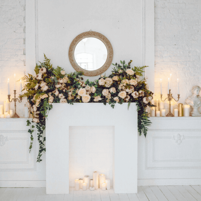 A white fireplace adorned with a lush garland of cream and pink flowers mixed with greenery. Above is a round, ornate mirror. Inside the fireplace are assorted lit candles, adding a warm glow to the room.