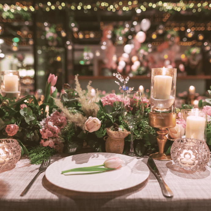 A beautifully set dinner table features a white plate with a pink tulip. The table is adorned with sparkling glass holders for rent, flameless candles, and a lush centerpiece of pink roses and greenery. Soft, warm lighting adds an elegant atmosphere.