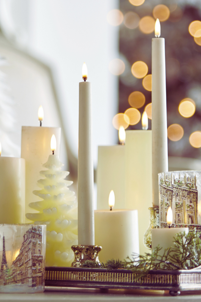 A cozy arrangement of lit candles in various shapes and sizes, including cylindrical and spiral tree forms, on a decorative tray with glass holders. Soft, warm glow and blurred bokeh lights in the background create a festive atmosphere.