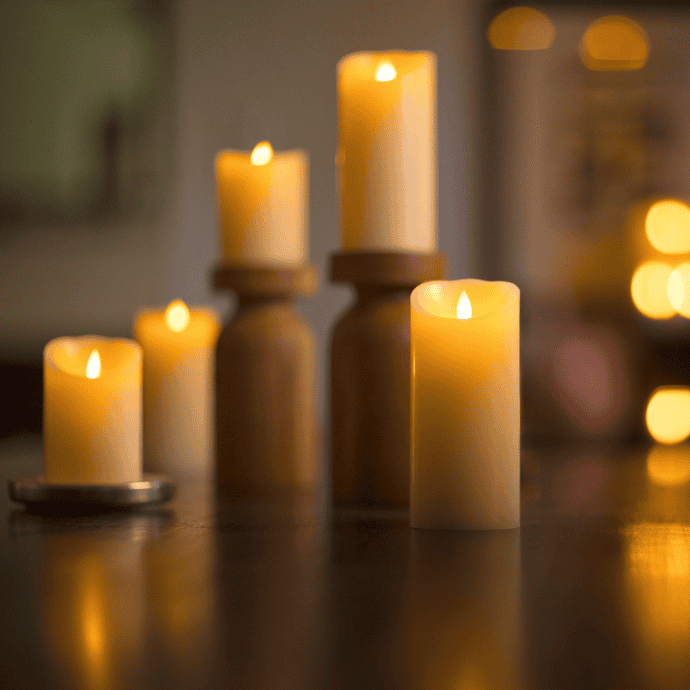 A group of lit candles of varying heights on a dark wooden surface. The candles have warm, glowing flames, creating a cozy and inviting atmosphere. The background is softly blurred with additional lights visible.