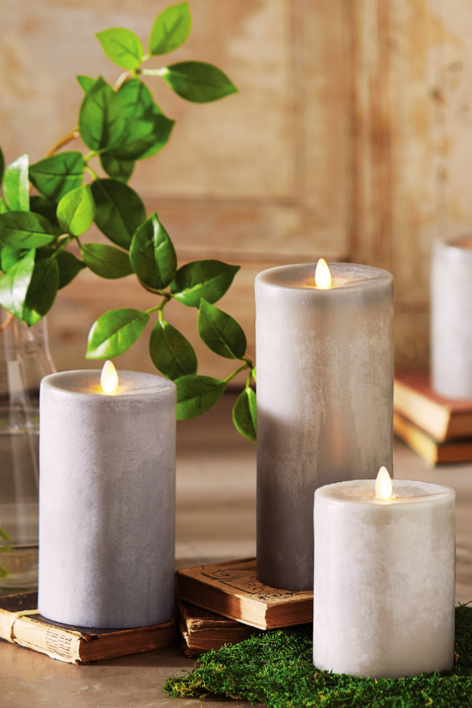 Three gray candles with flickering flames are arranged on vintage books and moss. A glass vase with green leafy branches sits in the background, creating a serene and cozy ambiance.