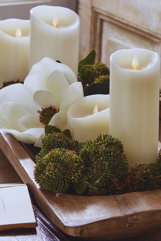 A wooden tray holds several lit white candles surrounded by green foliage and white magnolia flowers, creating a cozy and elegant display on a table.