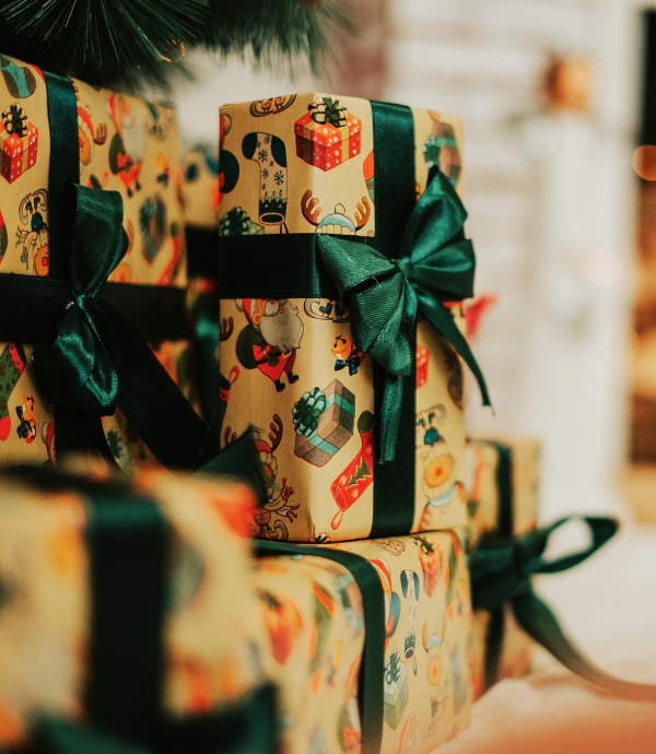 Wrapped gifts adorned with colorful paper featuring festive patterns and tied with dark green satin ribbons are stacked in a cozy, warmly lit setting, suggesting early Christmas gift planning is well underway.