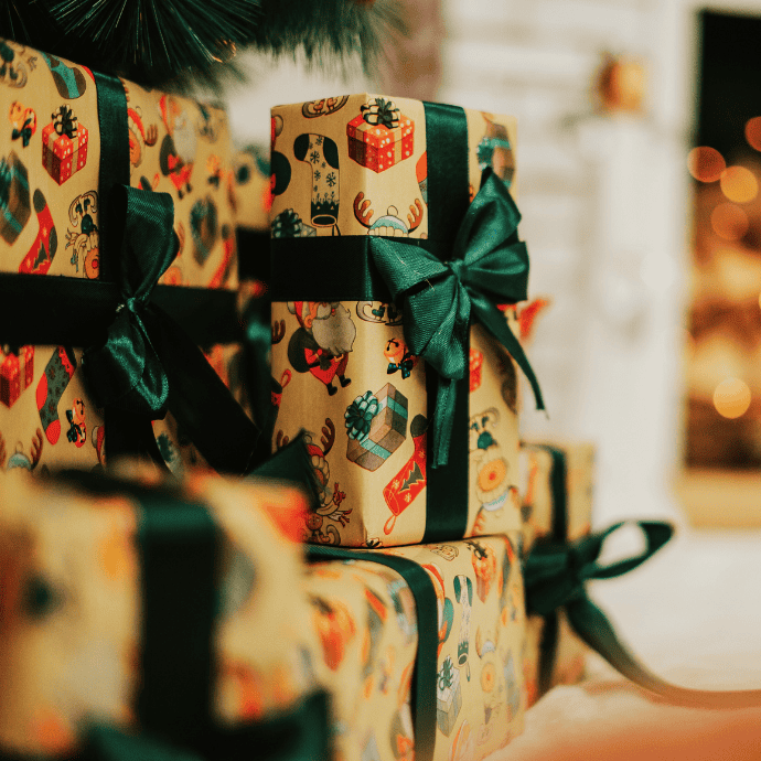 Wrapped gifts adorned with colorful paper featuring festive patterns and tied with dark green satin ribbons are stacked in a cozy, warmly lit setting, suggesting early Christmas gift planning is well underway.