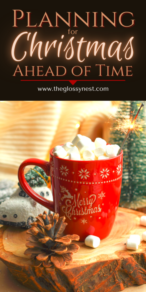 A red mug with "Merry Christmas" sits on a wooden slab, filled with marshmallows and a sparkler on top. Pine cones and a cozy scarf are nearby. Text above reads, "Planning for Christmas Ahead of Time." Website: www.theglossynest.com.