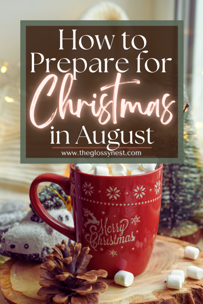 A red mug with "Merry Christmas" on it filled with marshmallows sits on a wooden surface. Snowy decorations and pine cones surround it. A text overlay reads, "How to Prepare for Christmas in August" with theglossynest.com URL.