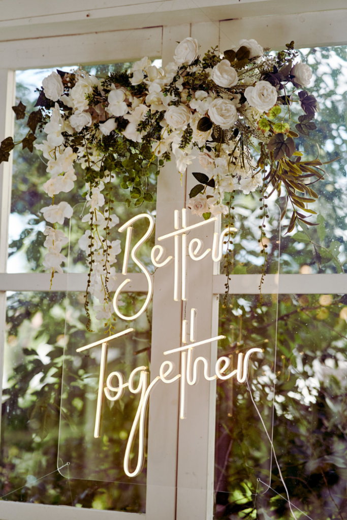 A window adorned with white flowers and greenery showcases an illuminated sign reading "Better Together," a perfect wedding signage idea. Sunlight filters through the glass, enhancing the floral arrangement's details.