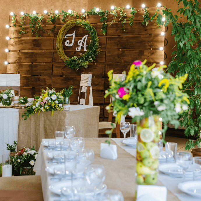 A decorated wedding reception area showcases a wooden backdrop with a circular wreath boasting the initials "J A", complemented by elegant neon signs with the couple's last name. The tables are adorned with white settings, candles, and vibrant floral arrangements, including a tall vase of fruits and flowers.