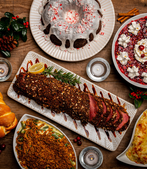 A festive table setting features sliced roast beef with seasonings, a frosted bundt cake, a red gelatin dessert topped with whipped cream, a green bean casserole, braided bread, and a cheesy dish. Decorated with candles and holly.