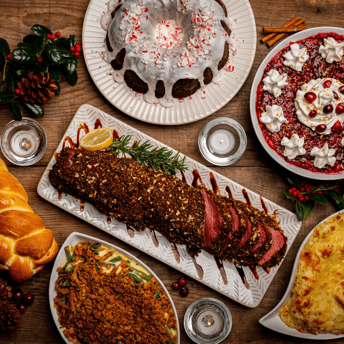 A festive table setting features sliced roast beef with seasonings, a frosted bundt cake, a red gelatin dessert topped with whipped cream, a green bean casserole, braided bread, and a cheesy dish. Decorated with candles and holly.