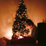 A person sits on the floor in pajamas, reading a book in front of a decorated, lit Christmas tree. A dog plush toy is beside them, adding to the cozy holiday atmosphere.