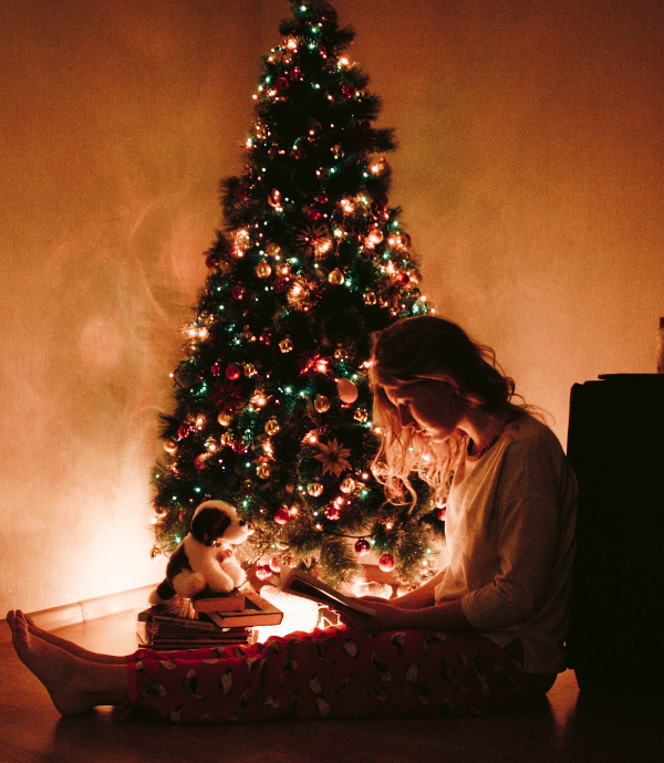 A person sits on the floor in pajamas, reading a book in front of a decorated, lit Christmas tree. A dog plush toy is beside them, adding to the cozy holiday atmosphere.