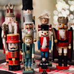 A collection of colorful nutcracker figures displayed on a table with a red and white checkered cloth. The nutcrackers vary in size and design, featuring soldiers, a king, and a drummer, set against a softly blurred background of lights.