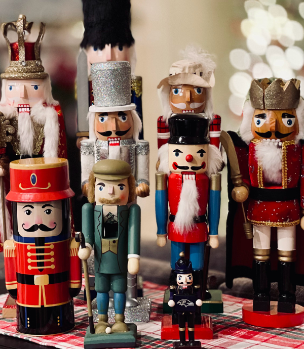 A collection of colorful nutcracker figures displayed on a table with a red and white checkered cloth. The nutcrackers vary in size and design, featuring soldiers, a king, and a drummer, set against a softly blurred background of lights.