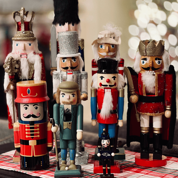 A collection of colorful nutcracker figures displayed on a table with a red and white checkered cloth. The nutcrackers vary in size and design, featuring soldiers, a king, and a drummer, set against a softly blurred background of lights.