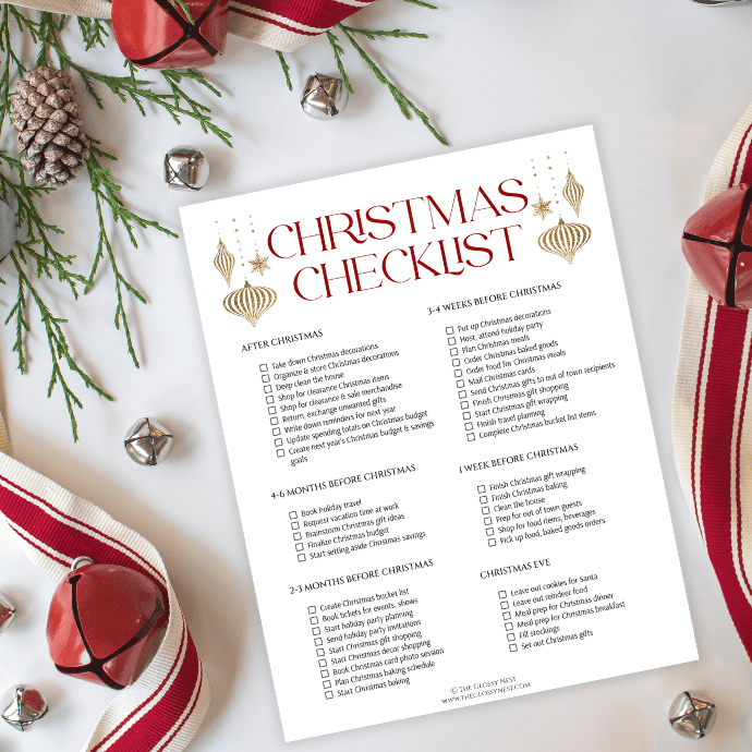 A flat lay image featuring a Christmas checklist on a white sheet, surrounded by red jingle bells, pinecones, and red and white ribbon. The checklist includes tasks and decorative elements like gold ornaments and stars.