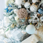 A close-up of a beautifully decorated Christmas tree adorned with silver, blue, and white ornaments. Several wrapped gifts in matching colors sit underneath the tree, creating a festive and elegant scene.