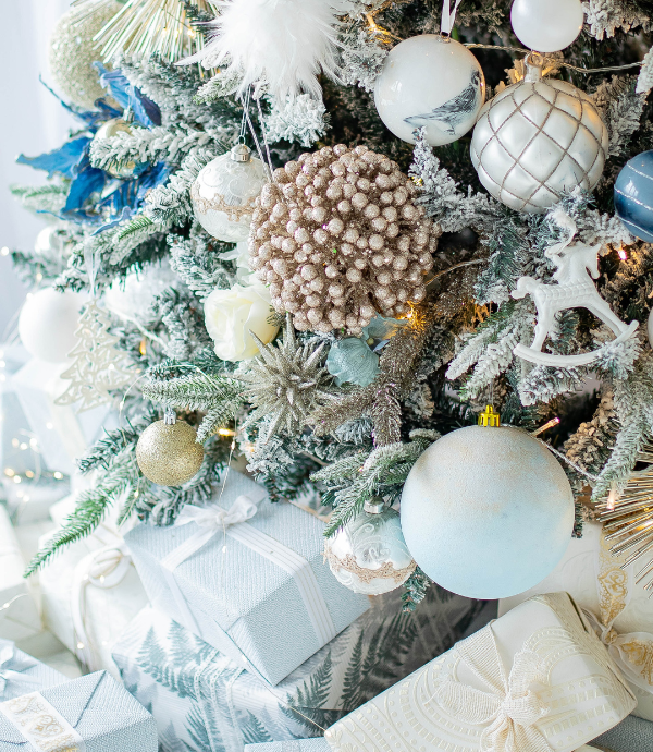 A close-up of a beautifully decorated Christmas tree adorned with silver, blue, and white ornaments. Several wrapped gifts in matching colors sit underneath the tree, creating a festive and elegant scene.