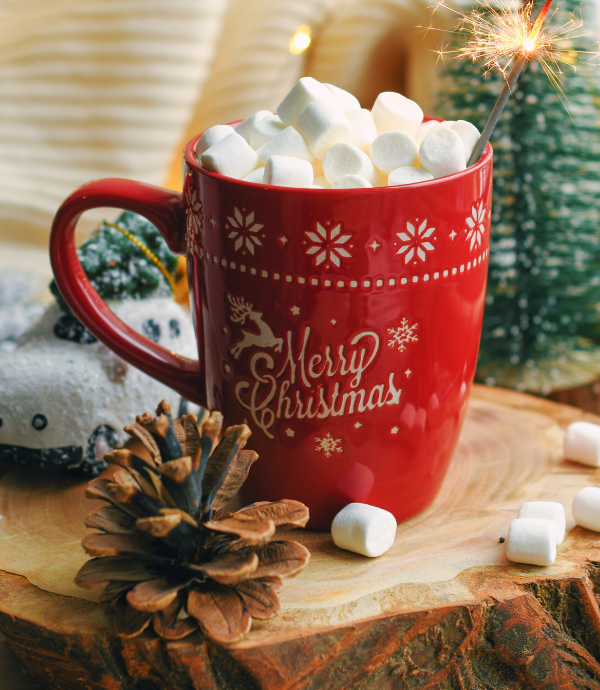 A red mug with "Merry Christmas" filled with hot chocolate and topped with marshmallows sits on a wooden surface. A lit sparkler is in the mug. A pine cone and snow-covered ornament are nearby, with soft lights in the background.