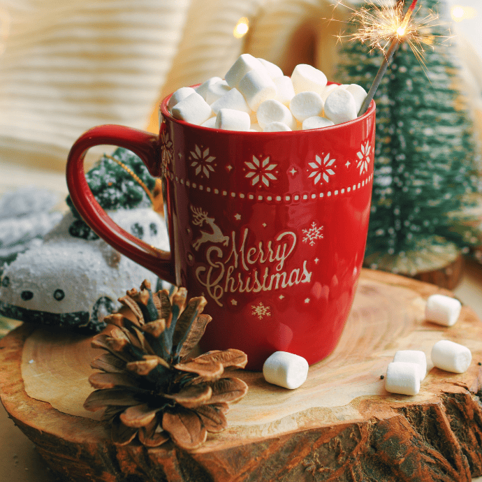 A red mug with "Merry Christmas" filled with hot chocolate and topped with marshmallows sits on a wooden surface. A lit sparkler is in the mug. A pine cone and snow-covered ornament are nearby, with soft lights in the background.