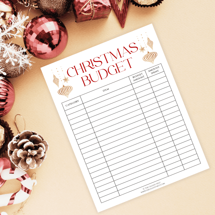 A Christmas budget planner sheet with columns for category, item, budget amount, and amount spent is placed on a beige surface. Surrounding it are festive decorations, including pink and gold ornaments, a pinecone, and a candy cane.