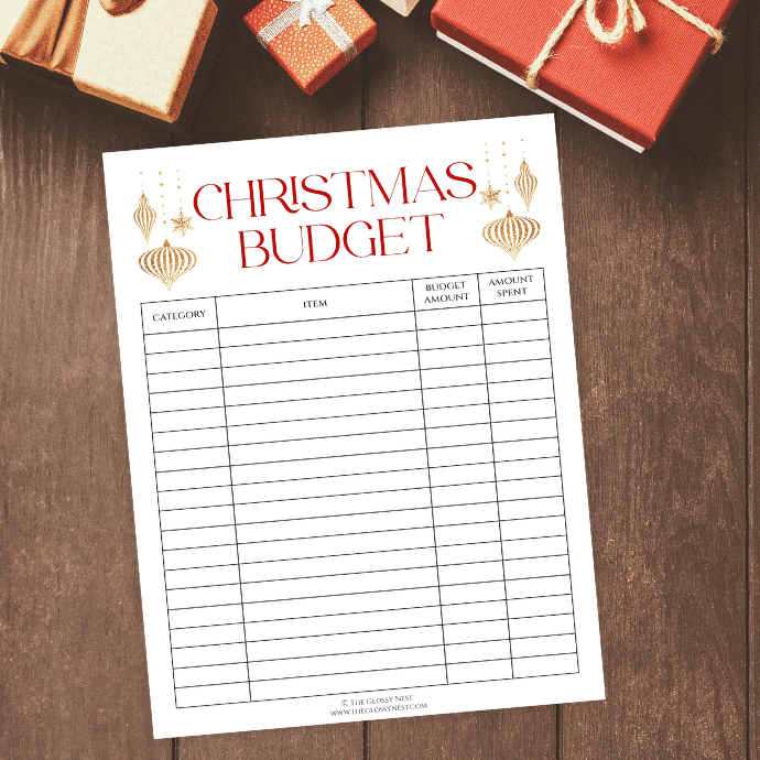 A Christmas budget planner on a wooden table with wrapped gifts nearby. The planner has spaces for category, item, budget amount, and amount spent, with decorative gold ornaments at the top.