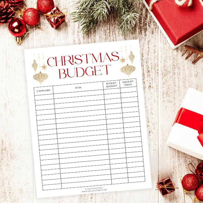 A Christmas budget planner is on a wooden table surrounded by festive decorations, including a wrapped gift, red and gold ornaments, and evergreen branches.