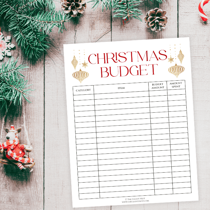 A Christmas budget planner sheet on a wooden table decorated with pine branches, pinecones, and a small reindeer ornament. The sheet has columns for category, item, budget amount, and amount spent.