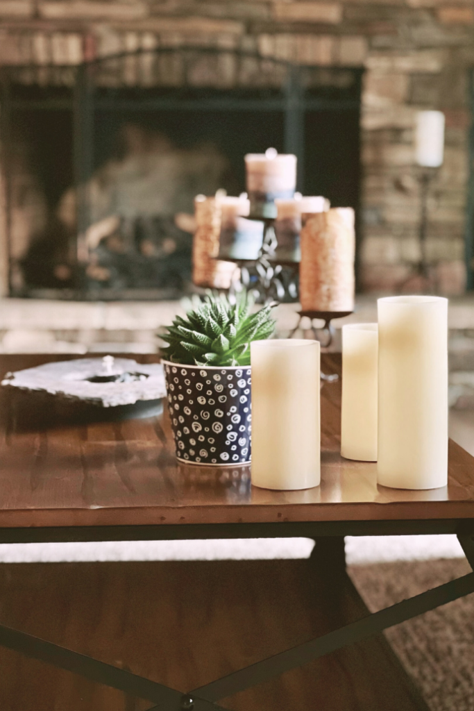 A cozy living room scene with a wooden coffee table featuring a small potted plant and several white candles. A stone fireplace with a candle display is in the background, creating a warm and inviting atmosphere.