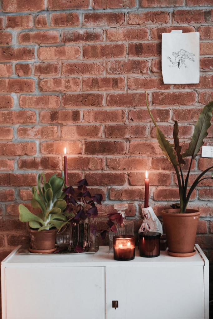 A cozy arrangement featuring lit candles, potted plants, and decorative items on a white cabinet against a brick wall. A small sketch is pinned to the wall above the cabinet.