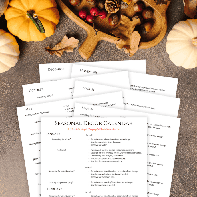 A collection of seasonal decor calendar pages spread out on a textured surface, surrounded by autumn-themed decorations, including pumpkins, acorns, leaves, and a wooden bowl with red berries and pinecones.