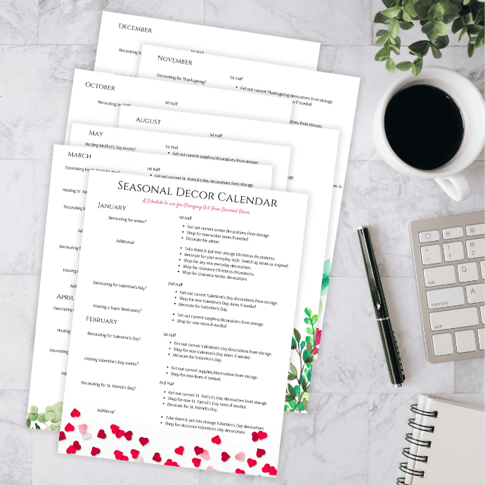 A desktop scene featuring a seasonal decor calendar with monthly pages spread out, a notepad, pen, keyboard, and a cup of coffee. Decorative elements include floral and heart designs on the pages.