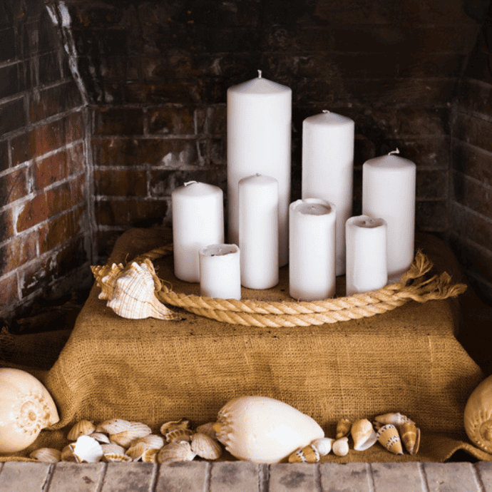 A collection of different-sized white candles arranged on a burlap-covered surface, surrounded by a thick rope. Seashells are scattered around the base, and the setting is inside a brick fireplace.