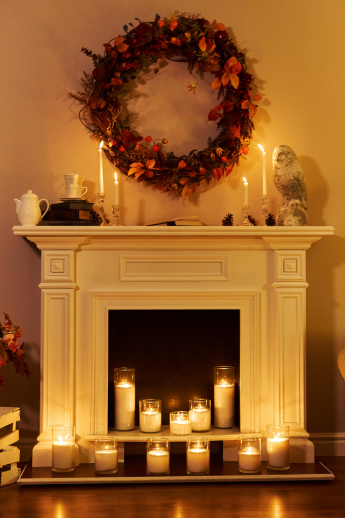 A cozy living room features a white fireplace adorned with a large autumn-themed wreath and lit candles. Various candles are arranged on the mantel and inside the fireplace, creating a warm, inviting atmosphere.