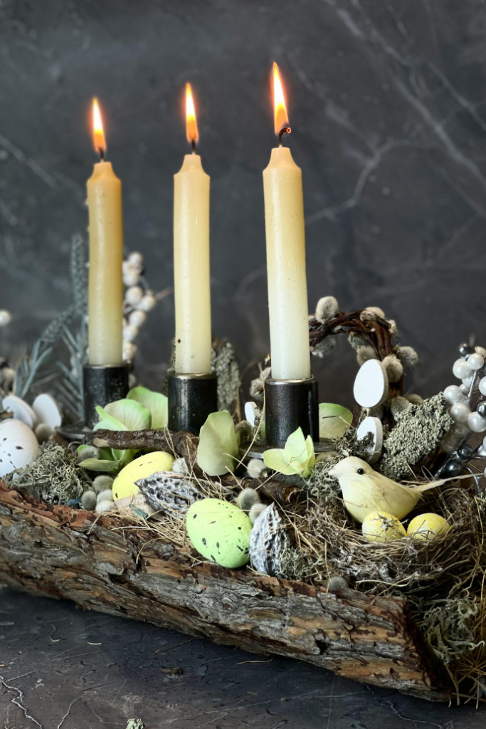 Three lit candles emit a soft glow from a rustic wooden holder, surrounded by decorative elements like speckled eggs, small nests, a yellow bird figurine, and various greenery on a dark marble background—perfect Easter candle ideas.