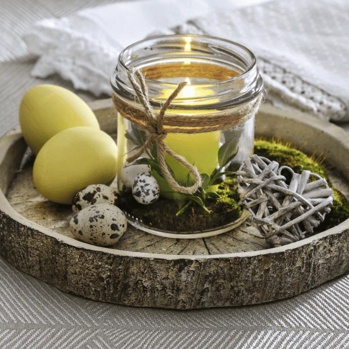 A rustic wooden tray holds a lit candle, casting a soft glow from its glass jar adorned with twine and greenery. Beside it, yellow eggs, speckled quail eggs, and a wicker heart rest on a mossy bed. In the background, a white cloth completes this charming Easter candle idea.