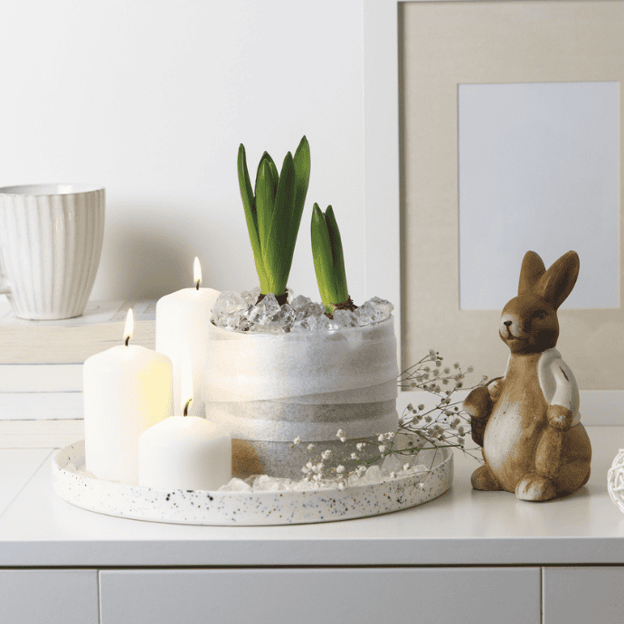 A white shelf featuring Easter candle ideas with lit candles on a speckled tray, a gray plant pot with green sprouts, and a small ceramic bunny figurine. A stack of books sits beside an empty picture frame, embodying cozy and minimalist decor.