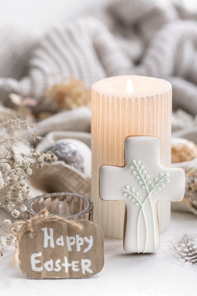 A lit candle casts a soft glow over a cross-shaped cookie with floral icing, nestled on a textured surface. A small tag reading "Happy Easter" leans against the candle, while dried flowers and cozy gray fabric create a blurred backdrop, perfect for Easter candle ideas.