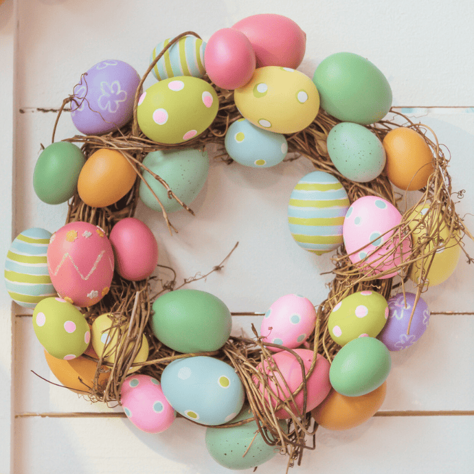 A wreath made of colorful Easter eggs and twigs is hanging on a white wall, serving as perfect Easter wreath ideas. The eggs feature patterns like polka dots, stripes, and flowers in pastel shades of pink, blue, yellow, green, and orange—ideal for a colorful front door display.