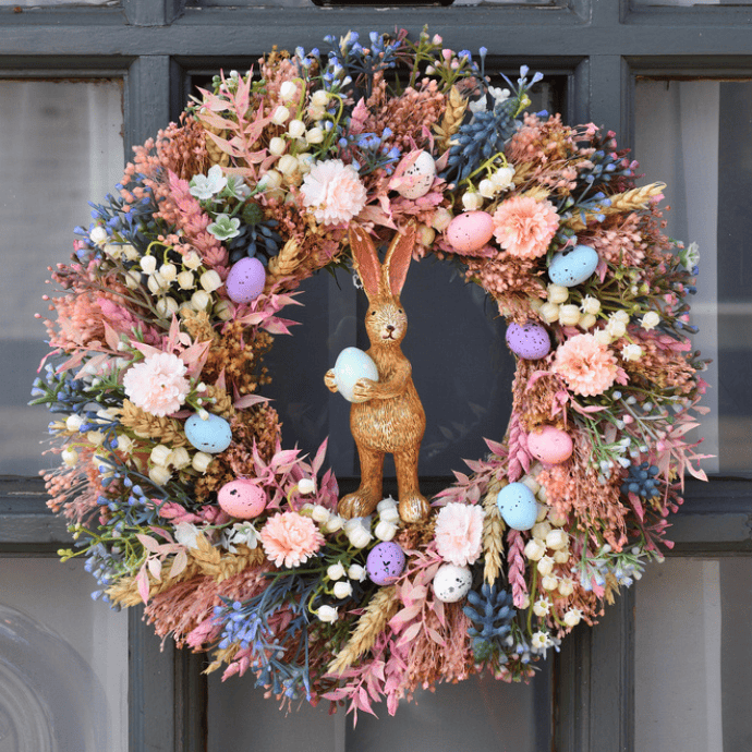 A decorative Easter wreath on the front door showcases a variety of colorful pastel flowers and speckled eggs. At its center, a rabbit holds a blue egg, with pink, purple, and blue elements adding to the festive, springtime theme. Perfect for those seeking colorful front door inspiration.