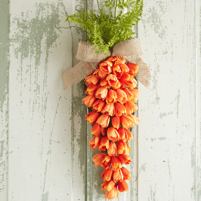 A colorful front door display features a decorative arrangement resembling a carrot, made of orange tulip flowers and green ferns, tied with a burlap bow. This charming piece offers unique Easter wreath ideas against a rustic, peeling white and green wooden background.