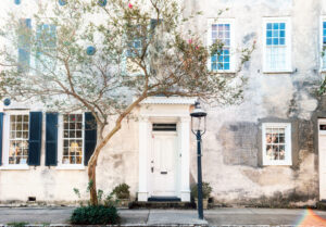 A historic, weathered white building features three windows with black shutters and a central white door. A tree with sparse leaves grows in front, and a classic black street lamp stands on the sidewalk. Subtle sunlight creates a gentle lens flare.